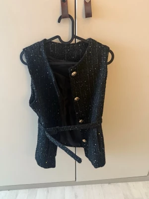 Svart glittrig väst med bälte - Stilren svart väst med glittriga inslag och knappar framtill. Västen har ett matchande bälte i midjan och är ärmlös med rak passform. Perfekt för att lyfta en outfit med lite extra bling.