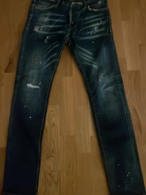 Mörkblå jeans med slitningar och färgstänk, dsq2 jeans äkta - Snygga mörkblå jeans med slitningar och färgstänk för en trendig och avslappnad look. Jeansen har klassisk femficksmodell och raka ben. Perfekta för dig som gillar en lite mer edgy stil.