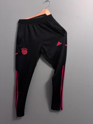 Adidas Bayern München träningsset - Snyggt träningsset från Adidas med Bayern München-logga. By soparna är svarta med röda detaljer och smala ben, jackan är vit med röda ränder och dragkedja. Perfekt för fotbollsträning eller sportig stil. Materialet är polyester.
