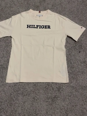 Vit Hilfiger t-shirt i bomull - Snygg vit t-shirt från Tommy Hilfiger med klassisk logga i svart text på bröstet. T-shirten har korta ärmar och en liten Hilfiger-flagga på ärmen. Tillverkad i mjuk bomull och har en rak passform som passar till det mesta.