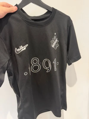 Svart AIK tröja Nike 1891 - Snygg svart AIK t-shirt från Nike med klubbmärke och texten '1891' på bröstet. Materialet är lätt och andas, perfekt för träning eller match. På ryggen står det 'SMOKINGLIRARE'. Klassisk passform och korta ärmar. Storlek S men passar M också!