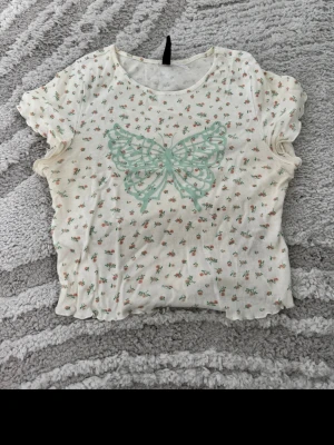 Blommig topp med fjäril från H&M Divided - Supersöt topp från H&M Divided med små blommor och en stor mintgrön fjäril på bröstet. Toppen är vit, har korta ärmar och är croppad med vågig kant. Perfekt för dig som gillar gulliga detaljer och en feminin stil.