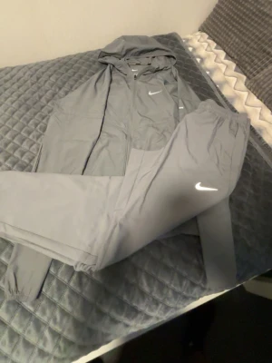 Grått Nike träningsset med huva - ÄKTA!!!!!!! Snyggt grått träningsset från Nike med matchande byxor och zip-hoodie. Jackan har huva och dragkedja, byxorna har resår i midjan och båda plaggen har den klassiska Nike-loggan. Perfekt för gymmet eller löprundan.