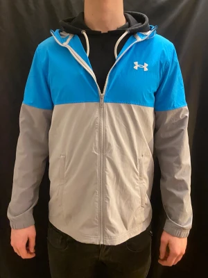 Windbreaker Under Armour - Sällsyn Under Armour vindjacka/windbreaker i blått och grått – en lätt, bekväm och funktionell jacka som skyddar bra mot vind och funkar perfekt både till träning och vardag. Utrustad med huva, hel dragkedja och två fickor för extra komfort och praktisk användning.  Snygg passform, lätt att matcha och i fint skick. Ett riktigt bra kap – passa på innan den försvinner. Kontakta mig vid intresse 👍