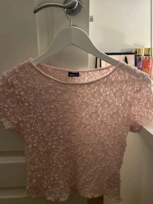  topp från Gina  - Söt ljusrosa topp från Gina i genombruten spets med blommigt mönster. Kortärmad och croppad modell med rund halsringning. Perfekt att styla med höga jeans eller kjol för en trendig look.