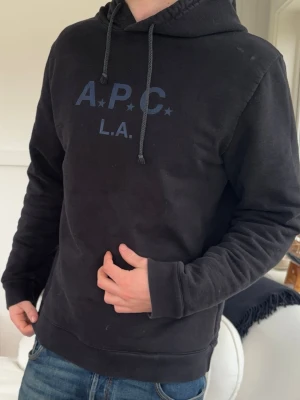 Svart hoodie från A.P.C. med tryck - Säljer en svart hoodie från A.P.C. med mörkblått tryck på bröstet där det står 'A.P.C. L.A.' och stjärndetaljer. Hoodien har huva med dragsko och ribbade muddar vid ärmar och nederkant. Perfekt för en chill och stilren look.