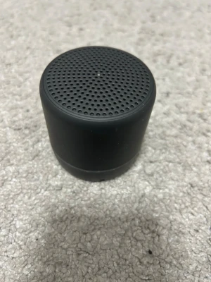 Svart bärbar mini-högtalare - Snygg och kompakt bärbar högtalare i svart med rundad cylinderform. Perfekt för att ta med sig överallt och spela musik trådlöst. Stilren design med perforerat galler på ovansidan för bra ljudspridning. 