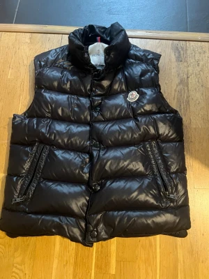 Svart dunväst från Moncler - Snygg svart dunväst från Moncler med glansig finish och klassisk logga på bröstet. Västen har hög krage, två dragkedjefickor och tryckknappar framtill. Insidan har ett unikt serietryck. Perfekt för lager-på-lager-stil under kyliga dagar.