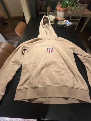 Beige hoodie från GANT med logga - Snygg beige hoodie från GANT med broderad logga och sköld på bröstet. Klassisk känguruficka framtill och huva. Mjuk bomullsblandning som är perfekt för en avslappnad stil.