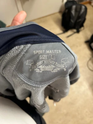 Gråblå vindjacka från Sport Master - Snygg gråblå vindjacka från Sport Master med marinblå detaljer över bröstet och dragkedja framtill. Jackan har hög krage, ribbade muddar och två fickor. Perfekt för dig som gillar sportig stil och vill ha en lätt jacka till vardagen.