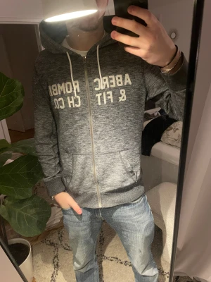 Grå hoodie från Abercrombie & Fitch - Snygg gråmelerad hoodie från Abercrombie & Fitch med dragkedja och vit logga framtill. Tröjan har huva med vita snören och ribbade muddar vid ärmslut och nederkant. Perfekt för en avslappnad och cool stil.