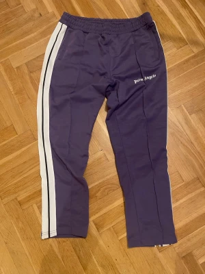 Palm Angels lila mjukisbyxor med ränder - Säljer ett par lila mjukisbyxor från Palm Angels med vita sidoränder och svart tunn linje. Byxorna har elastisk midja och logga broderad på vänster sida. Perfekta för en avslappnad och trendig look.