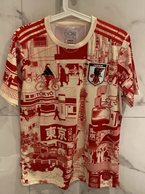 Adidas Japan Tokyo matchtröja röd/vit - Unik matchtröja från Adidas och Japan JFA i vitt och rött med detaljerade Tokyo-motiv över hela plagget. Korta ärmar, rund hals och officiellt JFA-märke på bröstet. Perfekt för dig som gillar streetstyle och sportmode med japansk touch. Är också nästan använd 