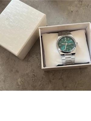 Silverfärgad klocka med grön urtavla - Stilren klocka från Poedagar med silverfärgad länk i metall och en grön urtavla. Klockan har dag- och datumvisning samt tydliga vita markeringar och visare. Klassisk design som passar till många olika stilar.