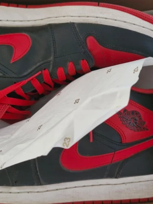 Nike Air Jordan 1 svart/röd sneakers - Säljer ett par Nike Air Jordan 1 sneakers i svart och rött skinn. Klassisk mid modell med röda snören, röda detaljer och ikoniska Air Jordan-loggan på sidan. Perfekta för dig som vill sticka ut med en tidlös och sportig look. Stl 46 men sitter som 44-45. Creease skydd ingår