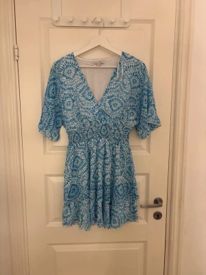 Blåvit omlottklänning med mönster - Kort klänning i blått och vitt med omlottmodell och elastisk midja. Klänningen har ett grafiskt mönster, v-ringad hals och korta ärmar. 