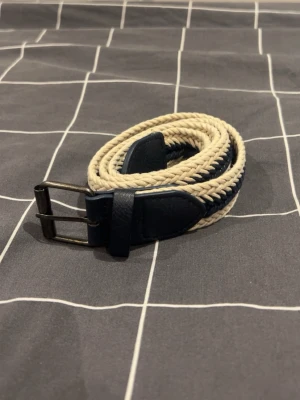 Flätat beige och blått bälte - Snyggt flätat bälte i beige och marinblått med detaljer i mörkblått läder och metallspänne. Perfekt accessoar för att lyfta din outfit med en stilren och avslappnad känsla. Passar väldigt snyggt till grisch stilen och den är 90 cm, den är helt oanvänd:) DM för mer info/ intresse
