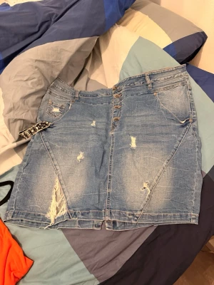 Blå jeanskjol med slitningar - Säljer en blå jeanskjol med slitna detaljer och knappar framtill. Kjolen har fickor både fram och bak samt en rå kant nertill. Perfekt för dig som gillar en avslappnad och trendig stil.