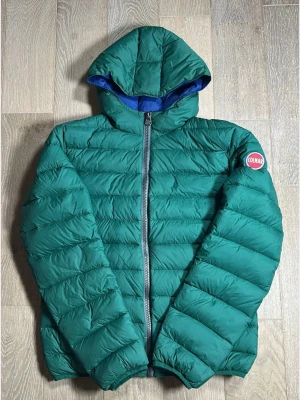 Grön dunjacka med huva från Colmar - Snygg grön dunjacka från Colmar med huva och blått innerfoder. Jackan har quiltad design, dragkedja framtill och Colmar-logga på ärmen. Perfekt för kalla dagar och enkel att matcha med olika outfits.