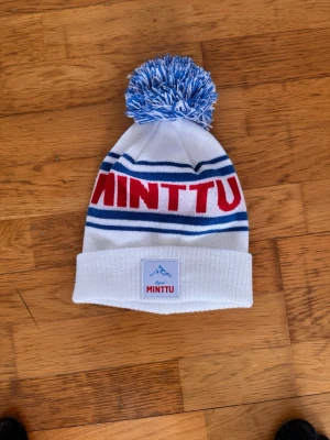 Minttu mössa med tofs - Säljer en vit och blå mössa från Minttu med röd text framtill och blåa ränder. Mössan har en stor blåvit tofs på toppen och vikt kant med Minttu-logga. Perfekt för kalla dagar och snygg till vinterlooken.