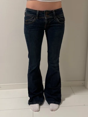 Mörkblå bootcut jeans lågmidjade - Snygga mörkblå jeans med bootcut-modell och låg midja. Klassisk femficksdesign och kontrastsömmar. 