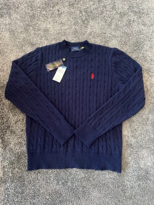 Mörkblå kabelstickad tröja Polo Ralph Lauren - Kabelstickad mörkblå tröja från Polo Ralph Lauren med rund halsringning och röd broderad logga på bröstet. Tröjan har ribbade muddar vid ärmslut och nederkant. Perfekt för dig som gillar klassisk och stilren design. Tröjan är helt ny med prislapp. Hör av dig vid funderingar!