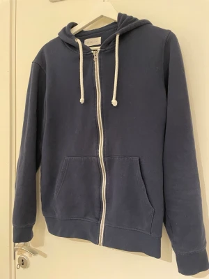 Marinblå hoodie med dragkedja Pier One - Snygg marinblå hoodie från Pier One med vit dragkedja och vita snören i huvan. Klassisk modell med kängurufickor framtill och mjukt bomullsmaterial. Perfekt för en avslappnad stil och enkel att matcha med jeans eller joggers.