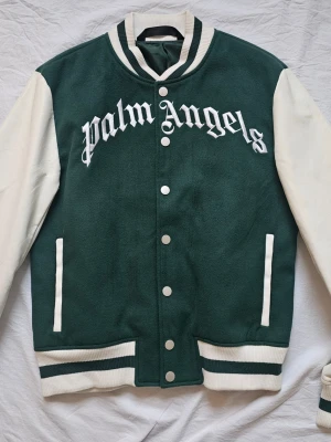 Palm Angels grön bomberjacka - Grön och vit bomberjacka från Palm Angels med stora broderade bokstäver framtill och en nallebrodyr på ryggen. Jackan har vita ärmar, ribbade muddar och knappar framtill. Klassisk varsity-look med kontrastfärgade detaljer. Aldrig använd!