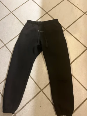 Svarta mjukisbyxor Essentials L - Svarta mjukisbyxor från Essentials med elastisk midja och snörning. Byxorna har en klassisk loose passform och är tillverkade i mjukt bomullsmaterial. Perfekta för en chill och avslappnad stil. Storleken är L men skulle säga att de sitter som storlek M