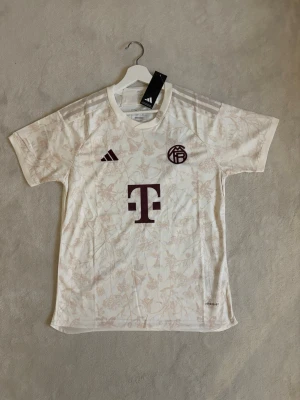 Bayern München tröja - Vit fotbollströja från Adidas för Bayern München med diskret blommigt mönster och vinröda detaljer. Korta ärmar, rund hals och klubbmärke samt Adidas-logga på bröstet. 