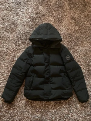 Svart dunjacka från Canada Goose - Snygg svart dunjacka från Canada Goose med huva och klassisk patch på ärmen. Jackan har en quiltad design, dragkedja framtill och två sidofickor. Perfekt för kalla dagar och riktigt varm tack vare dunfyllningen.