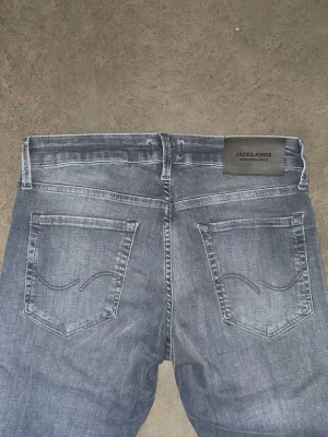 Grå slim jeans från Jack & Jones - Snygga grå slim jeans från Jack & Jones med klassisk femficksdesign och diskreta sömmar på bakfickorna. Jeansen har en smal passform och är tillverkade i mjuk bomullsdenim med lite stretch för extra komfort. De är helt nya och aldrig använda.    Jeansen är i storlek W28 L30