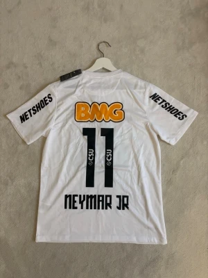 Neymar JR matchtröja  - Säljer en vit Santos fotbollströja från Nike med Neymar JR och nummer 11 på ryggen. Tröjan har tryckta sponsorer i svart, orange och rött samt klubbmärke på bröstet. Tillverkad i lätt polyester med korta ärmar och rund hals.