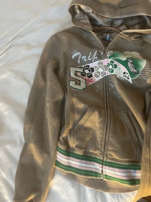 Y2K hoodie (vintage)med tryck och dragkedja - Säljer en brun hoodie från Triple Five Soul med dragkedja, stor huva och coolt tryck med siffran 5, blommor och fågel på bröstet. Ribbstickade muddar och midja med gröna, vita och rosa ränder. Mjuk bomullskänsla och kängurufickor framtill.