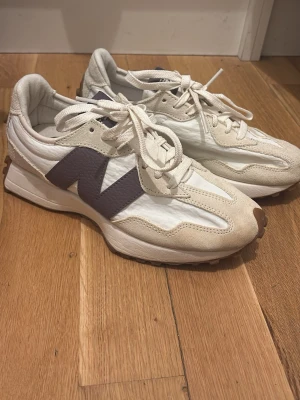 New Balance vita sneakers med lila detalj - Säljer ett par snygga vita New Balance sneakers med lila N-logga på sidan. Skorna har snörning, rund tå och en chunky sula med bruna detaljer baktill. Ovandelen är i en mix av mocka och syntetmaterial för en modern look. Använt bara en gång !