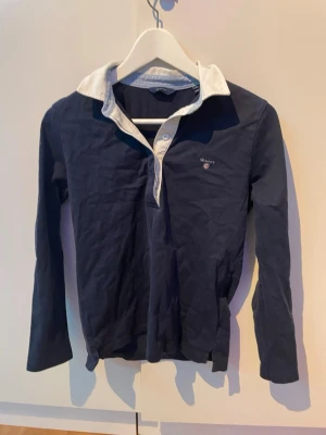 Mörkblå långärmad skjorta från Gant - Mörkblå långärmad skjorta från Gant med vit krage och knappslå. Klassisk logga broderad på bröstet. Tillverkad i mjuk bomull och har en stilren, normal passform. Perfekt för en clean och avslappnad look.