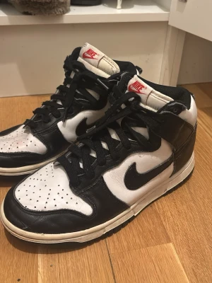 Nike Dunk High svartvita sneakers - Säljer ett par Nike Dunk High sneakers i svart och vitt läder med klassisk Swoosh på sidan. Skorna har högt skaft, svart snörning och vit sula med svarta detaljer. Tungan är vit med röd Nike-logga. Perfekta för dig som gillar streetwear och retrostil.