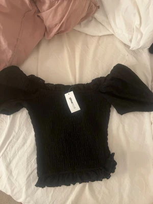 Svart offshoulder topp från gina - Trendig svart offshoulder topp från gina med puffiga korta ärmar och smockad kropp. Toppen har volangdetaljer vid nederkanten och är perfekt för en snygg och ungdomlig stil.