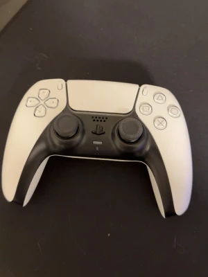 Sony DualSense PS5 handkontroll vit - Säljer en vit Sony DualSense handkontroll till PlayStation 5. Kontrollern har svart detalj runt styrspakarna och touchpad i mitten. Ergonomisk design med klassiska PlayStation-knappar och triggers. Perfekt för gaming på PS5.
