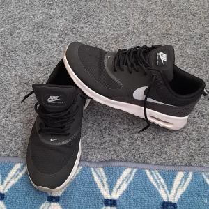 Svarta Nike Air Max Thea sneakers - Snygga svarta Nike Air Max Thea sneakers med vit sula och klassisk Nike-logga i vitt på sidan. Ovandelen är i mesh och syntet för en sportig look. Skorna har snörning och en låg profil, perfekt för en clean och stilren vibe.passar för smala fötter.