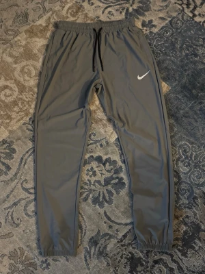 Gråa mjukisbyxor från Nike - Snygga grå mjukisbyxor från Nike med svart snörning i midjan och klassisk vit Nike-logga på benet. Byxorna har elastisk midja och benslut för skön passform. Perfekta för chill eller träning, tillverkade i mjukt syntetmaterial.