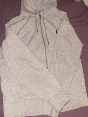Ljusgrå hoodie från Polo Ralph Lauren - Snygg ljusgrå hoodie från Polo Ralph Lauren med dragkedja framtill och klassisk huva med snören. Broderad logga i mörkblått på bröstet. Tillverkad i mjuk bomullsblandning och har ribbade muddar vid ärmslut och nederkant. Perfekt för en avslappnad stil.