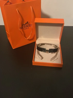 Svart HERMÈ4S S Clic H armband RESSERVERAD KÖP EJ - Stilrent svart Clic H armband från Hermè4s i silverfärgad metall med emaljdetaljer. Armbandet har den ikoniska H-formade låset och levereras i originalask och påse. Perfekt accessoar för att lyfta din outfit med lyxig känsla.