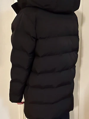 Svart lång dunjacka - Säljer en svart, lång dunjacka med huva och breda quiltade paneler. Jackan har dragkedja framtill och två stora fickor på sidorna. Perfekt för kalla dagar och riktigt skön att bära. Passar dig som gillar en klassisk och stilren vinterlook.