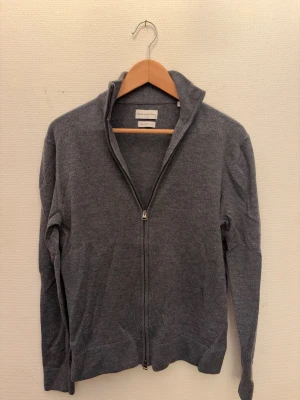 Grå fullzip tröja i merinoull - Mjuk och stilren merinoull ziptröja från These Glory Days i storlek M. Perfekt till att bara slänga på sig över en T-shirt för att hålla sig varm och få ett extra lager. Mycket bra skick förutom den defekt som syns på sista bilden. Skriv om ni har någots fler frågor!