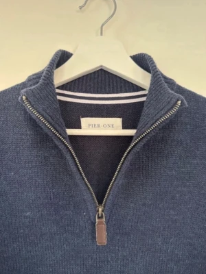 Mörkblå halvzip-tröja från Pier One - Mörkblå stickad tröja från Pier One med halvzip och hög krage. Tröjan har långa ärmar, ribbade muddar och en stilren look som passar perfekt till jeans. Materialet är mjukt och bekvämt, troligen bomull eller bomullsblandning.