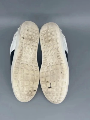 Valentino Garavani vita sneakers - Snygga vita sneakers från Valentino Garavani med en bred svart remsa över mitten och klassiska nitar på hälen. Skorna har rund tå, snörning och är tillverkade i skinn för en lyxig känsla. Perfekta för dig som gillar stilrena och exklusiva sneakers. Perfekt nu till när vintern börjar ta slut och de börjar bli vår.