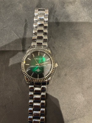 Silvrig klocka från Grealy - Stilren klocka från Grealy med rund svart urtavla och silvrigt armband i metall. soppskuren boett och tydliga indexmarkeringar. Klassisk design som passar till många olika stilar. Länkat armband ger en modern känsla.