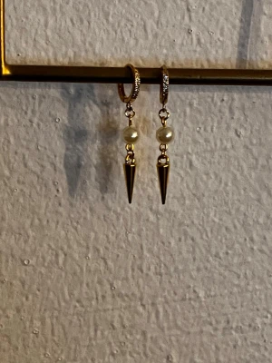 Guldörhängen med pärla och spets - Säljer ett par trendiga örhängen i guldfärg med små vita pärlor och hängande spetsformade detaljer. Örhängena har en rund creolbas och cool kontrast mellan den mjuka pärlan och den spetsiga metallformen. Perfekta för dig som gillar edgy accessoarer.