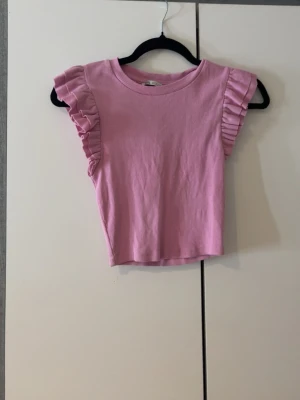 Rosa croppad topp med volang från Zara - Supersöt rosa croppad topp från Zara med volangdetaljer på ärmarna. Toppen är ribbad och har en rund halsringning. Perfekt för dig som vill ha en feminin och trendig look.